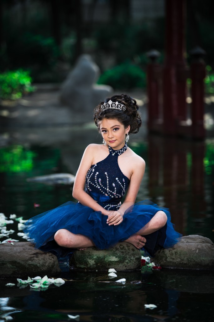 A young girl in a sparkling blue dress and a tiara sits on a rock in a serene pond surrounded by greenery and flower petals.